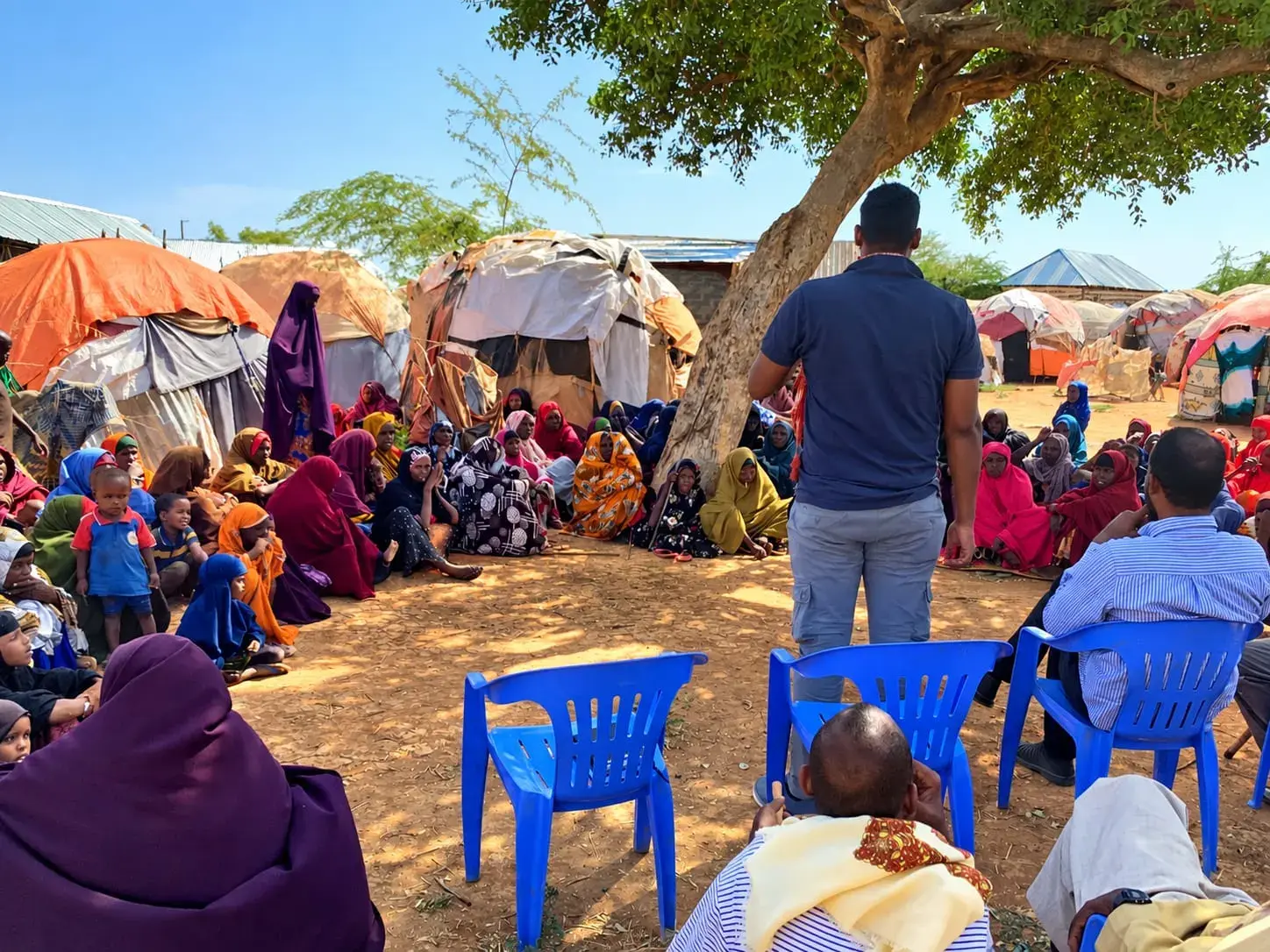 A man stands in the shade of a tree speaking to a large gathering of women and children seated on the ground, with tarpaulin shelters behind them.