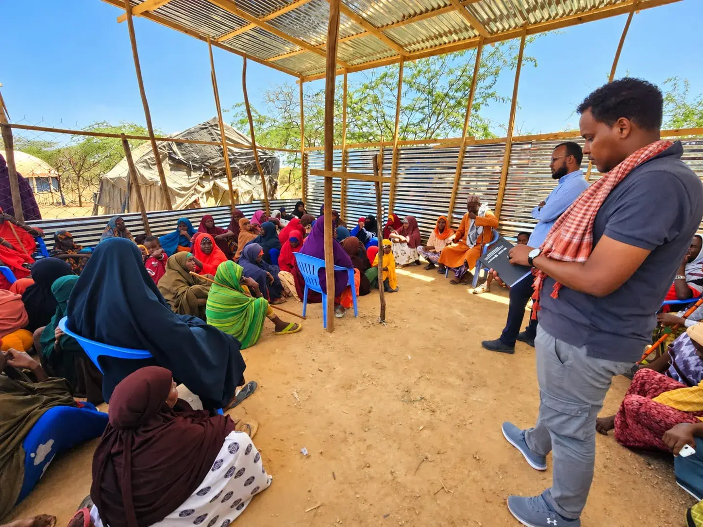 Two men stand in an open timber-and-sheet-metal shelter, one holding a folder, speaking with women seated on the ground and on chairs.