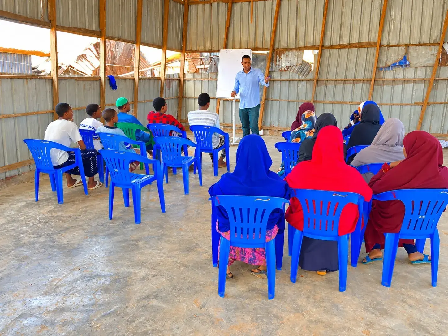 A facilitator at a whiteboard leads a session for young men and women seated on blue plastic chairs inside a sheet-metal shelter.