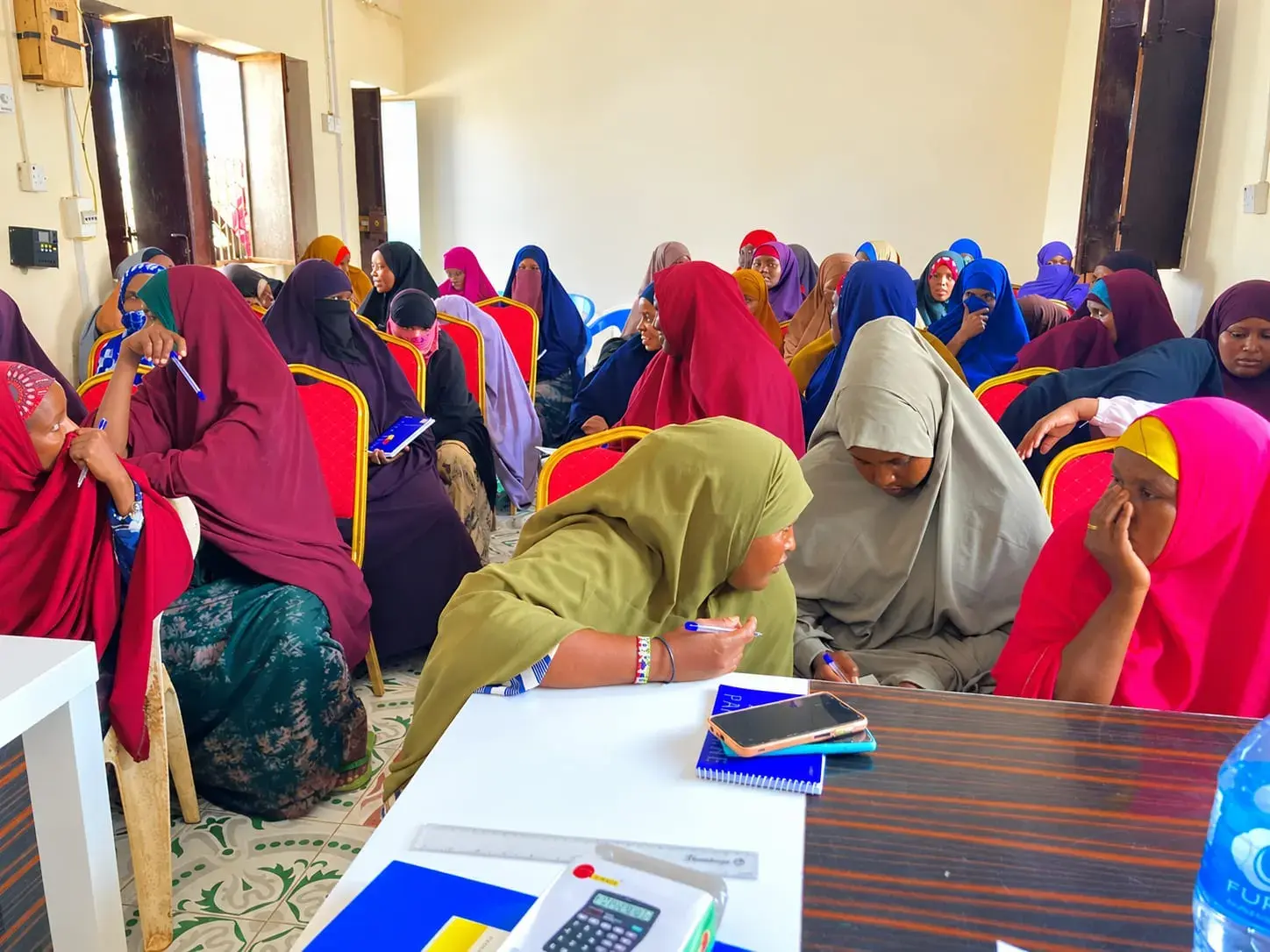 Women seated in a classroom at a workshop, some writing in notebooks, while two women at the front talk together.