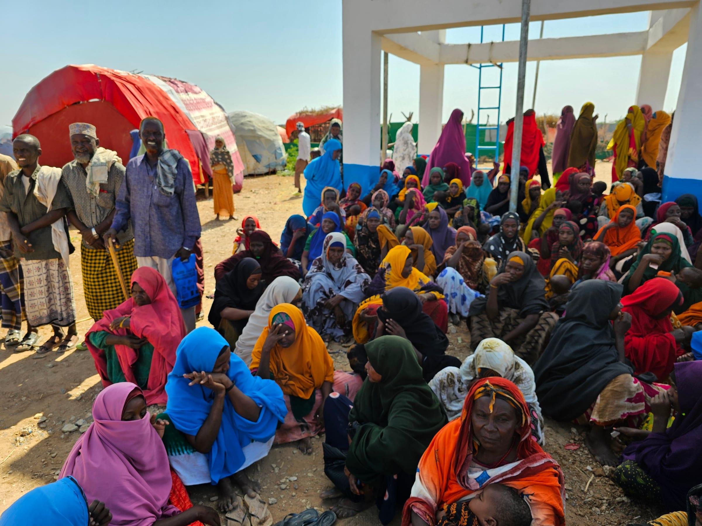 A large group of women, some holding small children, seated on the ground beside a white building, with older men standing to the left.