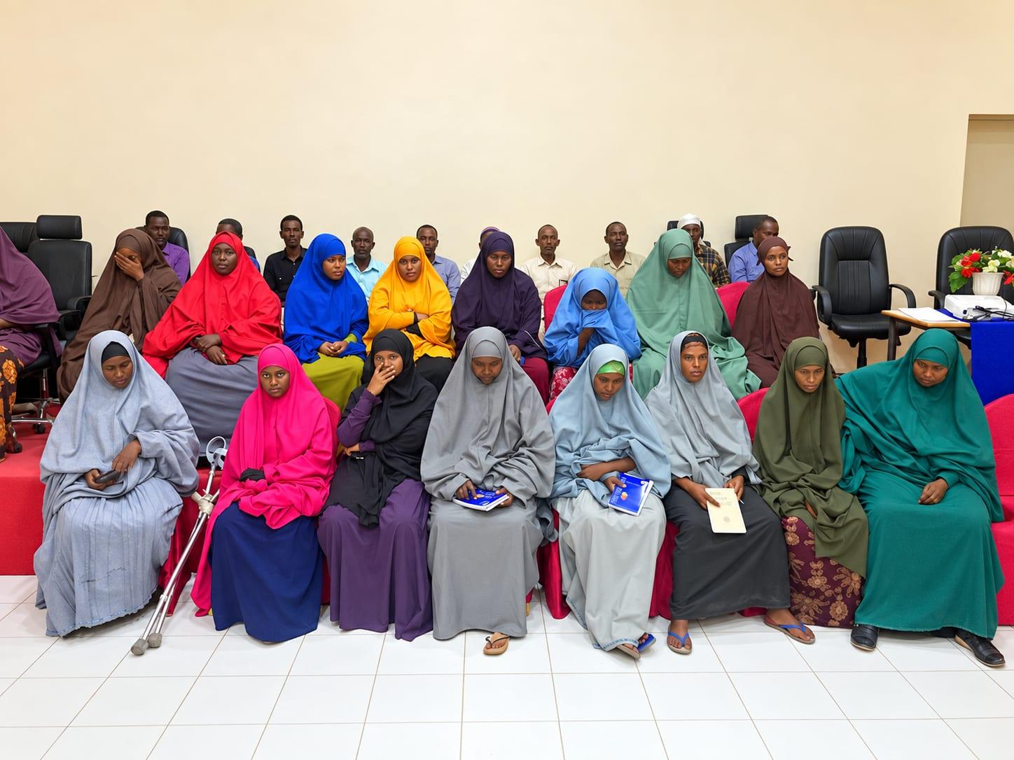 Women in brightly coloured hijabs seated in two rows at a training session, some holding notebooks, with men seated behind them.